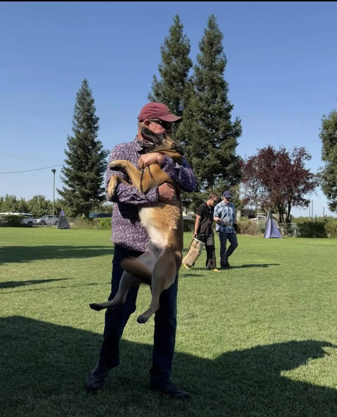 Joel Monroe hugs Cash, a Belgian Malinois, lifting him off the ground on a grassy training field on a sunny day