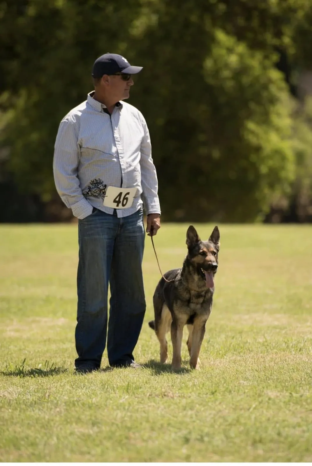 Joel Monroe stands on a sunny field wearing a number 46 bib, holding a German Shepherd on a loose leash at his side