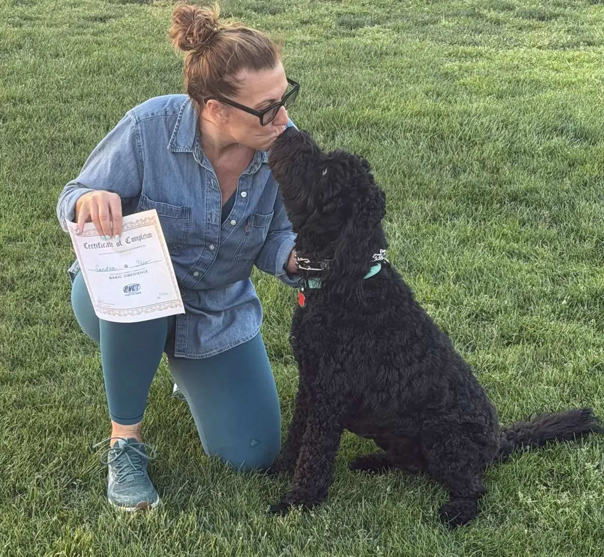 Woman kneeling on grass kisses her black curly-coated dog while holding a Basic Obedience certificate of completion