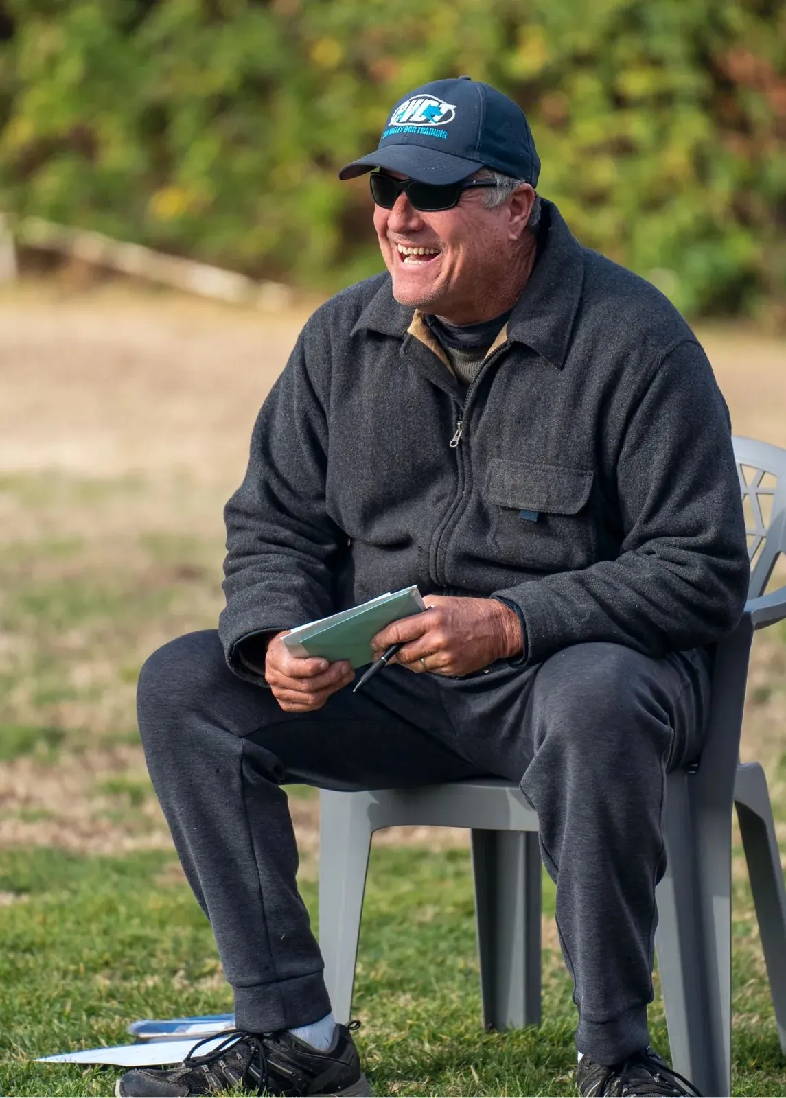 Joel Monroe laughs while sitting in a plastic chair on a field, holding a notepad and pen, wearing a BVDT cap